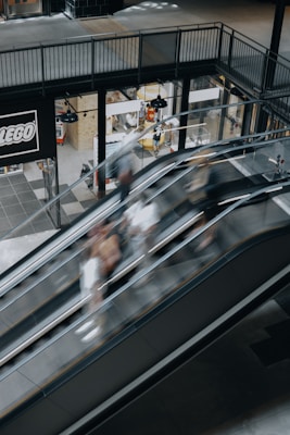 A shopping center setting with escalators showing motion blur of people moving up and down. The surrounding area includes a shop with a prominent LEGO sign, displaying various products through large windows. Industrial-style lighting hangs from the ceiling, and the flooring is a mix of tiles in neutral colors.