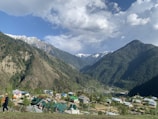 A picturesque valley surrounded by majestic mountains covered in dense forest. Snow-capped peaks are visible in the distance under a partly cloudy sky. Below, a small village with colorful rooftops nestles in the greenery, with a person walking in traditional attire in the foreground.