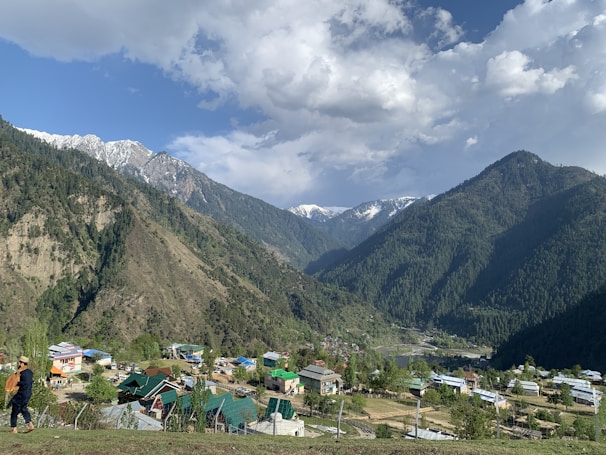 A picturesque valley surrounded by majestic mountains covered in dense forest. Snow-capped peaks are visible in the distance under a partly cloudy sky. Below, a small village with colorful rooftops nestles in the greenery, with a person walking in traditional attire in the foreground.