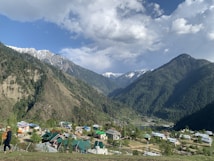 A picturesque valley surrounded by majestic mountains covered in dense forest. Snow-capped peaks are visible in the distance under a partly cloudy sky. Below, a small village with colorful rooftops nestles in the greenery, with a person walking in traditional attire in the foreground.