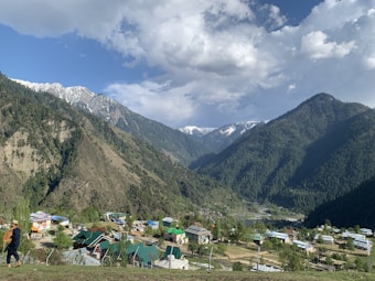 A picturesque valley surrounded by majestic mountains covered in dense forest. Snow-capped peaks are visible in the distance under a partly cloudy sky. Below, a small village with colorful rooftops nestles in the greenery, with a person walking in traditional attire in the foreground.