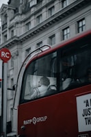A red bus with RATP Group branding is stopped near a building with classical architectural details. The bus has large windows revealing a passenger, who appears to be sitting near the window, visible in profile. The building behind the bus features ornate carvings and multiple windows. Part of a red and white bus stop sign is visible in the foreground.