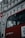 A red bus with RATP Group branding is stopped near a building with classical architectural details. The bus has large windows revealing a passenger, who appears to be sitting near the window, visible in profile. The building behind the bus features ornate carvings and multiple windows. Part of a red and white bus stop sign is visible in the foreground.