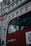 A red bus with RATP Group branding is stopped near a building with classical architectural details. The bus has large windows revealing a passenger, who appears to be sitting near the window, visible in profile. The building behind the bus features ornate carvings and multiple windows. Part of a red and white bus stop sign is visible in the foreground.