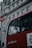 A red bus with RATP Group branding is stopped near a building with classical architectural details. The bus has large windows revealing a passenger, who appears to be sitting near the window, visible in profile. The building behind the bus features ornate carvings and multiple windows. Part of a red and white bus stop sign is visible in the foreground.