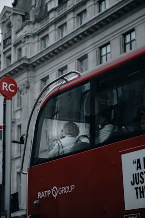 A red bus with RATP Group branding is stopped near a building with classical architectural details. The bus has large windows revealing a passenger, who appears to be sitting near the window, visible in profile. The building behind the bus features ornate carvings and multiple windows. Part of a red and white bus stop sign is visible in the foreground.