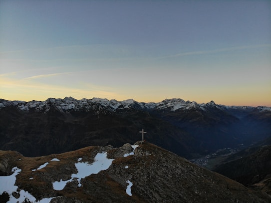 A serene mountain landscape with snow-capped peaks in the distance under a clear sky, transitioning from light to dark at sunset. A cross stands prominently on a rocky outcrop in the foreground, amidst patches of snow.