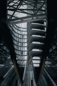 A high-contrast architectural scene featuring modern buildings with glass and metal structures. The image captures a series of escalators leading up to an overhanging architectural piece with curved and geometric design elements.