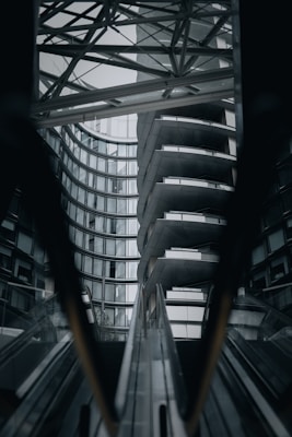 A high-contrast architectural scene featuring modern buildings with glass and metal structures. The image captures a series of escalators leading up to an overhanging architectural piece with curved and geometric design elements.