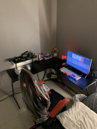 A gaming setup features a large corner desk with a computer monitor displaying the game Red Dead Redemption II. The desk holds a gaming chair still wrapped in protective plastic, a gaming keyboard with colorful backlighting, a game controller, and decorative items like a plush toy. A blanket and a pillow are on the side, suggesting a cozy setup.