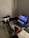 A gaming setup features a large corner desk with a computer monitor displaying the game Red Dead Redemption II. The desk holds a gaming chair still wrapped in protective plastic, a gaming keyboard with colorful backlighting, a game controller, and decorative items like a plush toy. A blanket and a pillow are on the side, suggesting a cozy setup.