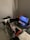 A gaming setup features a large corner desk with a computer monitor displaying the game Red Dead Redemption II. The desk holds a gaming chair still wrapped in protective plastic, a gaming keyboard with colorful backlighting, a game controller, and decorative items like a plush toy. A blanket and a pillow are on the side, suggesting a cozy setup.