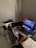 A gaming setup features a large corner desk with a computer monitor displaying the game Red Dead Redemption II. The desk holds a gaming chair still wrapped in protective plastic, a gaming keyboard with colorful backlighting, a game controller, and decorative items like a plush toy. A blanket and a pillow are on the side, suggesting a cozy setup.