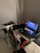 A gaming setup features a large corner desk with a computer monitor displaying the game Red Dead Redemption II. The desk holds a gaming chair still wrapped in protective plastic, a gaming keyboard with colorful backlighting, a game controller, and decorative items like a plush toy. A blanket and a pillow are on the side, suggesting a cozy setup.