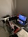 A gaming setup features a large corner desk with a computer monitor displaying the game Red Dead Redemption II. The desk holds a gaming chair still wrapped in protective plastic, a gaming keyboard with colorful backlighting, a game controller, and decorative items like a plush toy. A blanket and a pillow are on the side, suggesting a cozy setup.