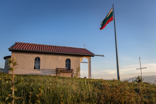 A small chapel with a red-tiled roof stands on a grassy hilltop. A large flagpole with a tricolor flag is nearby, and a metal cross is visible in the distance. The scene is bathed in warm, golden sunlight, typical of late afternoon.