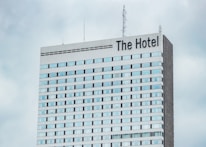A high-rise hotel building with multiple floors and a large sign reading 'The Hotel' at the top. The facade consists of light-colored panels and numerous windows. Antennas and communication equipment are visible on the roof, set against a cloudy sky.