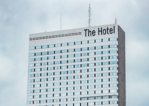 A high-rise hotel building with multiple floors and a large sign reading 'The Hotel' at the top. The facade consists of light-colored panels and numerous windows. Antennas and communication equipment are visible on the roof, set against a cloudy sky.