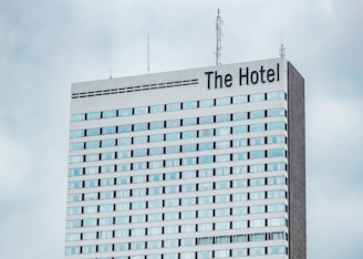 A high-rise hotel building with multiple floors and a large sign reading 'The Hotel' at the top. The facade consists of light-colored panels and numerous windows. Antennas and communication equipment are visible on the roof, set against a cloudy sky.
