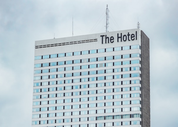 A high-rise hotel building with multiple floors and a large sign reading 'The Hotel' at the top. The facade consists of light-colored panels and numerous windows. Antennas and communication equipment are visible on the roof, set against a cloudy sky.