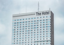 A high-rise hotel building with multiple floors and a large sign reading 'The Hotel' at the top. The facade consists of light-colored panels and numerous windows. Antennas and communication equipment are visible on the roof, set against a cloudy sky.