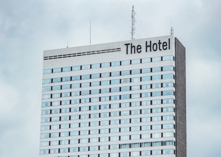 A high-rise hotel building with multiple floors and a large sign reading 'The Hotel' at the top. The facade consists of light-colored panels and numerous windows. Antennas and communication equipment are visible on the roof, set against a cloudy sky.