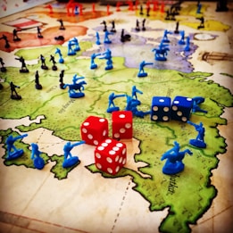 A strategic board game setup features a map of territories with plastic toy soldiers in blue and black positioned across various regions. Red and blue six-sided dice with white dots are scattered among the figures, indicating game moves or outcomes. The terrain colors indicate different zones, enhancing the gaming experience with visual aids.