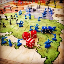 A strategic board game setup features a map of territories with plastic toy soldiers in blue and black positioned across various regions. Red and blue six-sided dice with white dots are scattered among the figures, indicating game moves or outcomes. The terrain colors indicate different zones, enhancing the gaming experience with visual aids.