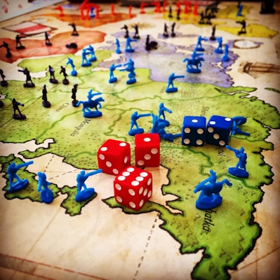 A strategic board game setup features a map of territories with plastic toy soldiers in blue and black positioned across various regions. Red and blue six-sided dice with white dots are scattered among the figures, indicating game moves or outcomes. The terrain colors indicate different zones, enhancing the gaming experience with visual aids.