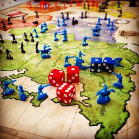 A strategic board game setup features a map of territories with plastic toy soldiers in blue and black positioned across various regions. Red and blue six-sided dice with white dots are scattered among the figures, indicating game moves or outcomes. The terrain colors indicate different zones, enhancing the gaming experience with visual aids.