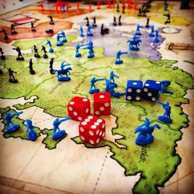 A strategic board game setup features a map of territories with plastic toy soldiers in blue and black positioned across various regions. Red and blue six-sided dice with white dots are scattered among the figures, indicating game moves or outcomes. The terrain colors indicate different zones, enhancing the gaming experience with visual aids.
