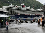 A large cruise ship is docked near a mountainous area with lush green hills. People are walking around on a damp pathway, some dressed in raincoats, indicating wet weather. A building with a sign that says 'TICKETS' is visible to the left, and a few people are standing near a colorful mascot on the right, adding a playful element to the scene. The atmosphere is lively with tourists exploring the area.