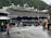 A large cruise ship is docked near a mountainous area with lush green hills. People are walking around on a damp pathway, some dressed in raincoats, indicating wet weather. A building with a sign that says 'TICKETS' is visible to the left, and a few people are standing near a colorful mascot on the right, adding a playful element to the scene. The atmosphere is lively with tourists exploring the area.