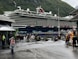 A large cruise ship is docked near a mountainous area with lush green hills. People are walking around on a damp pathway, some dressed in raincoats, indicating wet weather. A building with a sign that says 'TICKETS' is visible to the left, and a few people are standing near a colorful mascot on the right, adding a playful element to the scene. The atmosphere is lively with tourists exploring the area.