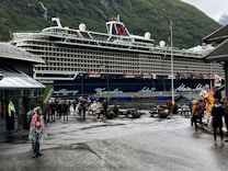 A large cruise ship is docked near a mountainous area with lush green hills. People are walking around on a damp pathway, some dressed in raincoats, indicating wet weather. A building with a sign that says 'TICKETS' is visible to the left, and a few people are standing near a colorful mascot on the right, adding a playful element to the scene. The atmosphere is lively with tourists exploring the area.