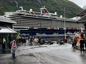A large cruise ship is docked near a mountainous area with lush green hills. People are walking around on a damp pathway, some dressed in raincoats, indicating wet weather. A building with a sign that says 'TICKETS' is visible to the left, and a few people are standing near a colorful mascot on the right, adding a playful element to the scene. The atmosphere is lively with tourists exploring the area.