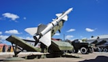A military missile is mounted on a launcher, aimed towards the sky. Surrounding the missile is military equipment, including trucks and containers. The scene is set outdoors under a bright blue sky with scattered fluffy clouds.