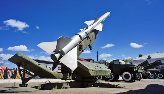 A military missile is mounted on a launcher, aimed towards the sky. Surrounding the missile is military equipment, including trucks and containers. The scene is set outdoors under a bright blue sky with scattered fluffy clouds.