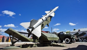 A military missile is mounted on a launcher, aimed towards the sky. Surrounding the missile is military equipment, including trucks and containers. The scene is set outdoors under a bright blue sky with scattered fluffy clouds.