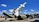A military missile is mounted on a launcher, aimed towards the sky. Surrounding the missile is military equipment, including trucks and containers. The scene is set outdoors under a bright blue sky with scattered fluffy clouds.