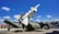 A military missile is mounted on a launcher, aimed towards the sky. Surrounding the missile is military equipment, including trucks and containers. The scene is set outdoors under a bright blue sky with scattered fluffy clouds.