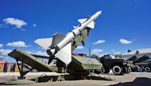 A military missile is mounted on a launcher, aimed towards the sky. Surrounding the missile is military equipment, including trucks and containers. The scene is set outdoors under a bright blue sky with scattered fluffy clouds.