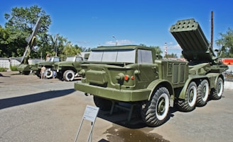 Several military vehicles and missile launch systems are displayed outdoors in a museum-like setting. The vehicles are painted in army green and are positioned on a concrete surface with informational plaques in front of them. In the background, there are trees and utility poles under a clear blue sky.