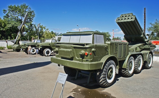Several military vehicles and missile launch systems are displayed outdoors in a museum-like setting. The vehicles are painted in army green and are positioned on a concrete surface with informational plaques in front of them. In the background, there are trees and utility poles under a clear blue sky.