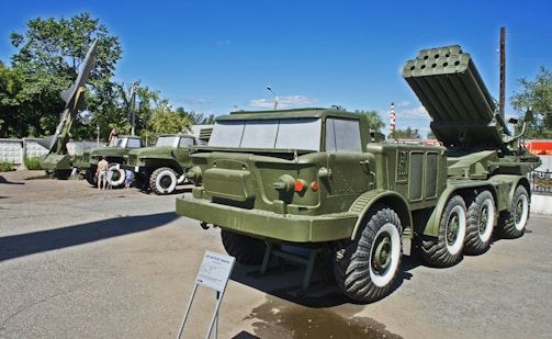 Several military vehicles and missile launch systems are displayed outdoors in a museum-like setting. The vehicles are painted in army green and are positioned on a concrete surface with informational plaques in front of them. In the background, there are trees and utility poles under a clear blue sky.