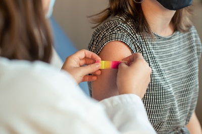 A person is applying a colorful band-aid to another person's upper arm. The individual receiving the band-aid is wearing a checkered sleeveless top and a black face mask.