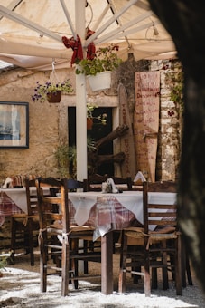 An outdoor dining area with several wooden chairs and tables covered with white tablecloths is shown. The tables are arranged in a cozy setting under a large canopy that provides shade. Flower pots hang from the canopy structure, adding a touch of greenery. There is a menu board with handwritten items displayed next to the entrance of a rustic stone building. The scene suggests an inviting and quaint restaurant or cafe.