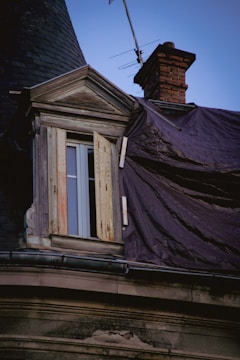 A close-up view of an old building's rooftop featuring a weathered window with partially open shutters. The roof is covered with worn shingles, and a plastic tarp is draped across part of it. A brick chimney and a television antenna are visible against the evening sky.