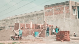 A construction site with workers engaged in building activities. The partially constructed structure is made of red bricks. Several workers are scattered around, one pushing a wheelbarrow loaded with sand, another climbing a wooden ladder. In the background, a large building with a rough, cemented exterior stands. Piles of materials like bricks and sand are visible, along with construction tools and equipment.