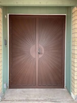 A large, double brown metal door with a sunburst design pattern originating from a central circular point where the door handles are located. The door is framed by a green wooden structure and two beige brick columns on each side.